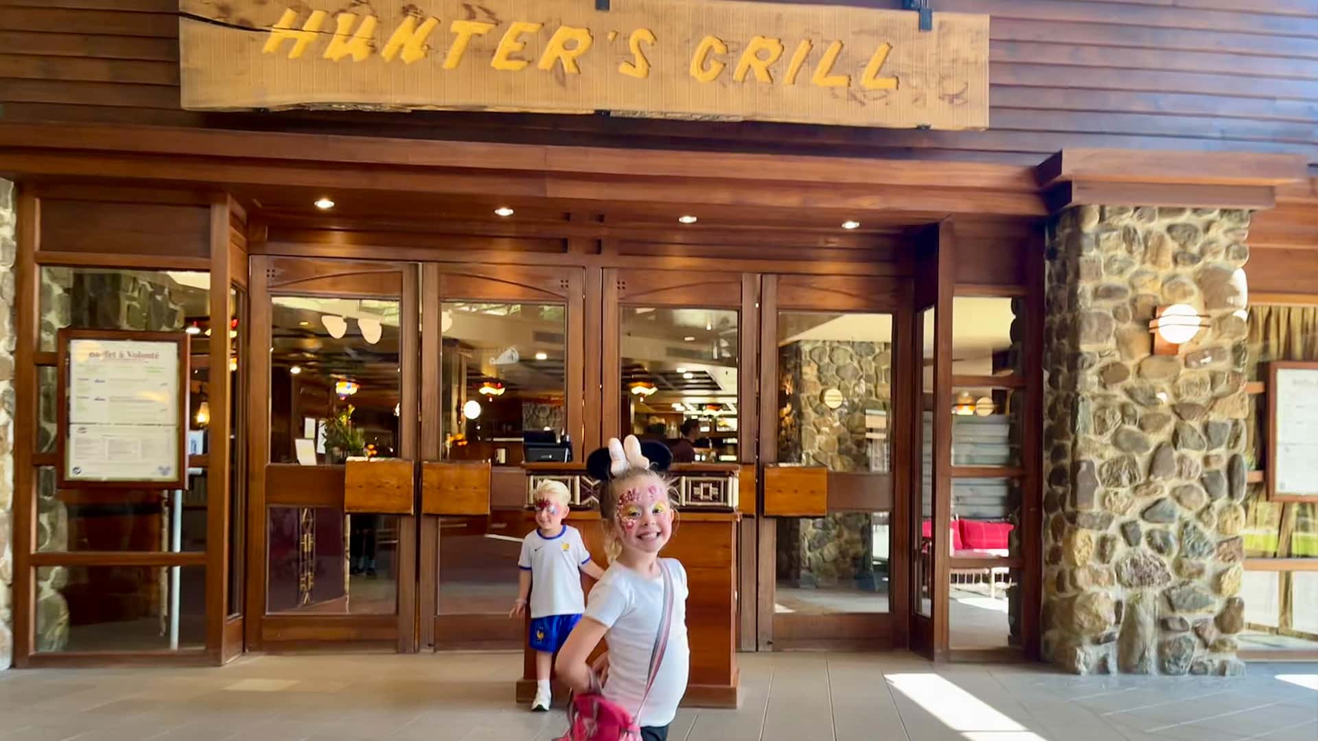 Lola en Jimmy kijken lachend in de camera voor de ingang van Hunter's Grill